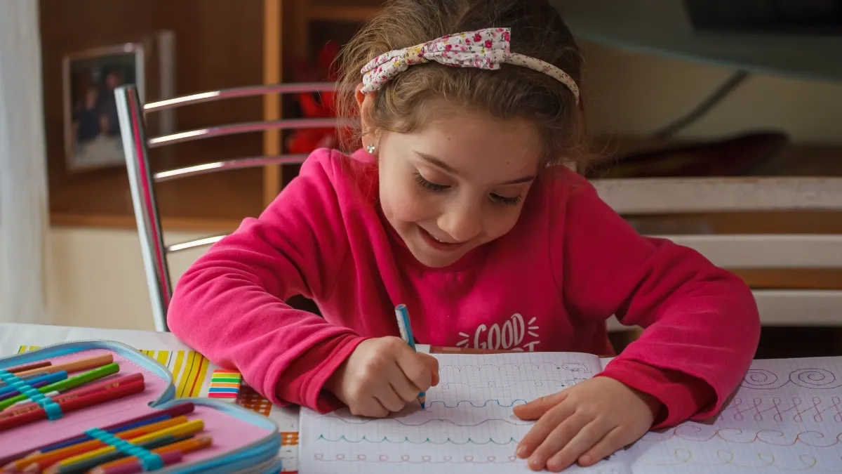 a girl child sitting at a table with a notebook and pencils